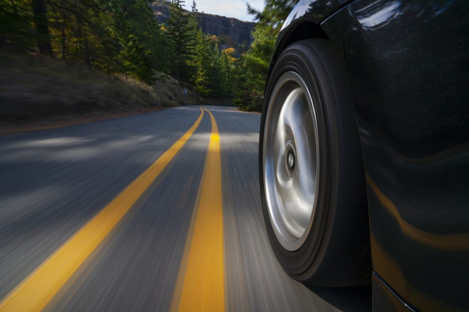 Close-up of a tire on a black car driving down a tree-lined street