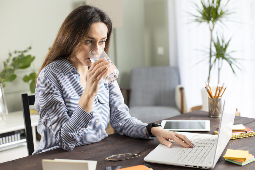 Young business woman working at home and drink glass of water. She drinking water while working at the laptop computer. Brown hair busy freelancer woman working at computer and connecting to the internet