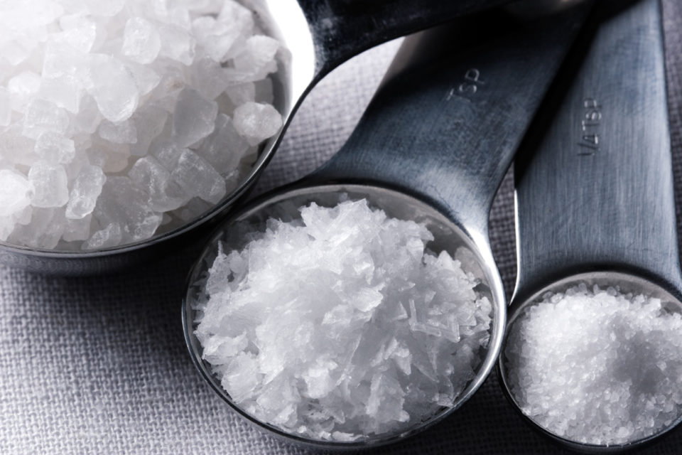 Close-up of three different types of salt in three measuring spoons. Kosher salt is on the left in a tablespoon, Maldon sea salt flakes are in the middle in a teaspoon, and fine sea slat is on the right in a quarter-teaspoon. 