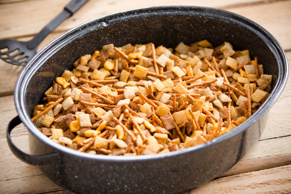 This is a shot of a party mix containing cereal, peanuts and pretzels in a roaster pan on an old wooden table.