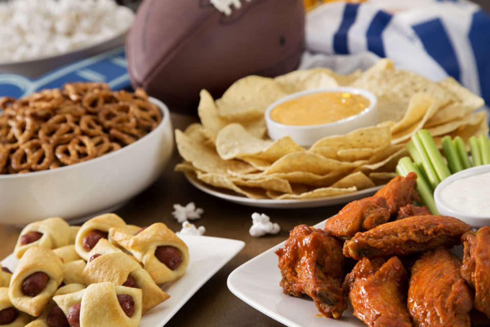 Tailgate party spread with hot wings, pig in a blanket, nachos, pretzels, and popcorn with football and a football jersey.