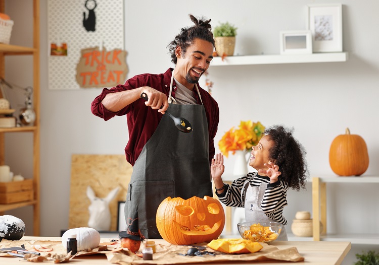 Smiling african american parent father removing pulp from ripe pumpkin while carving jack o lantern with little son for Halloween celebration at home in kitchen and looking at each other with smile