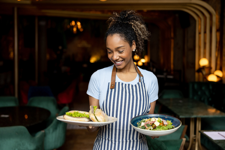 Portrait of a beautiful African American waitress serving food at a restaurant and smiling while carrying plates