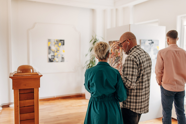 Diverse visitors in modern art gallery looking at paintings. mature couple looking at paintings and listening to audio guide on headphones.