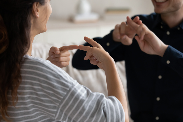 Young positive couple sit on sofa at home communicating using sign language, friends talking indoor showing fingers gestures, having fun, leading pleasant conversation, people with disability concept