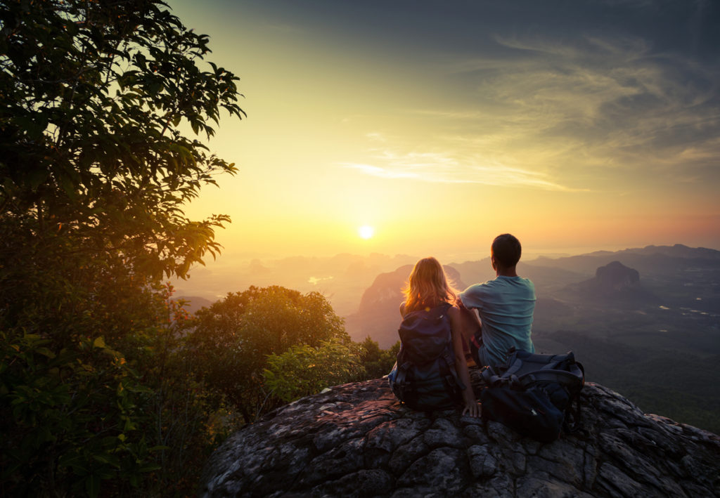 couple sitting on a cliff, enjoying a scenic mountain view with a the sun setting in the distance.