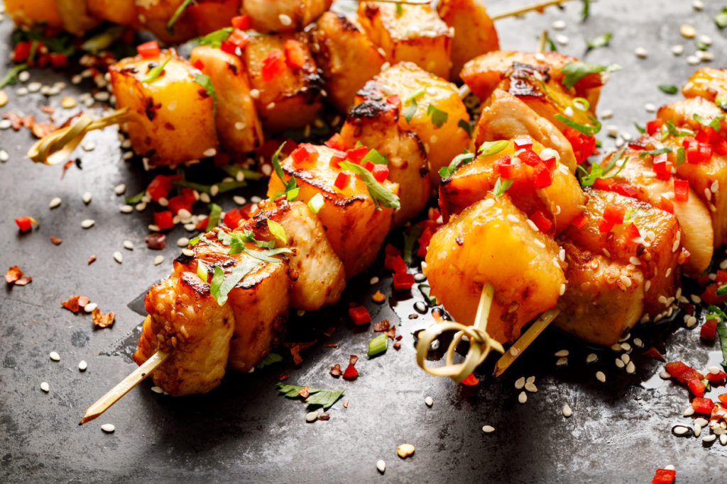 Grilled skewers with pineapple fruit and chicken meat sprinkled with sesame seeds, chilli pepper and fresh herbs on a black background