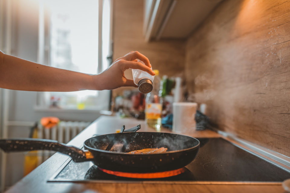 woman Adding salt. Back light to salt from salt shaker. Close up. Young Caucasian lady standing in the kitchen while adding salt to the meal in frying pan with chicken breast.