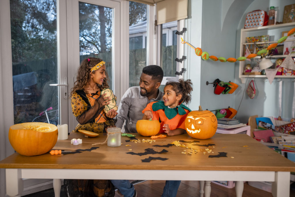 A man and his daughters sitting at their kitchen table at home in North East England during halloween, designing and carving pumpkins. One girl is showing her father and sister a butternut squash that she has designed for the halloween season.