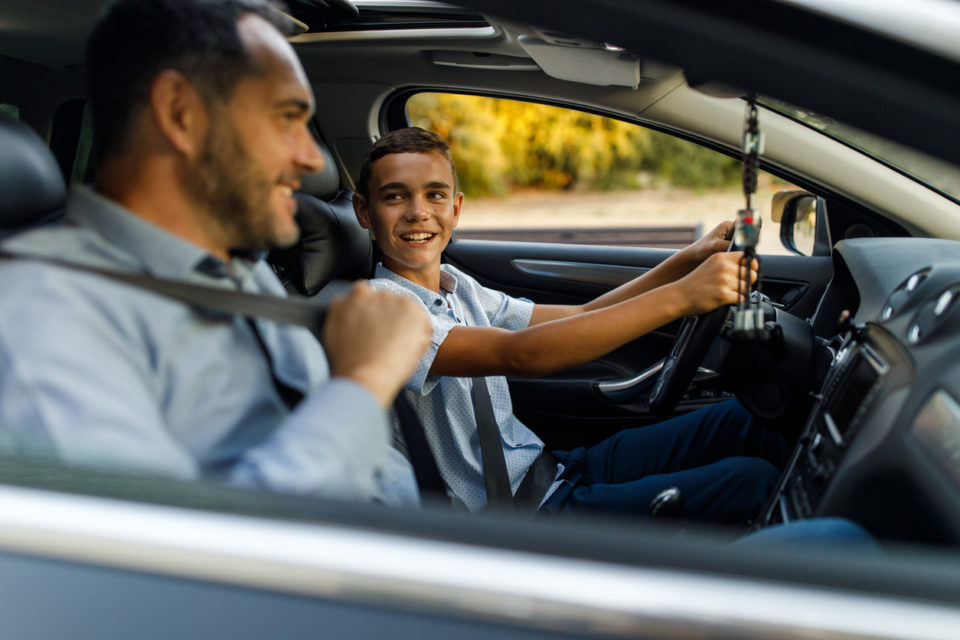 Selective focus shot of cheerful mid adult man sitting in passenger’s seat and putting on seatbelt when teaching his teenage son to drive.